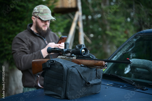 Bild auf Leinwand Hunter preparing scoped rifle on truck hood while checking phone near wooded hun