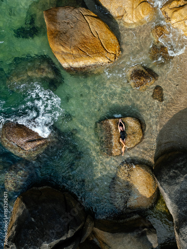 Aerial top view of woman lying on rock surrounded by turquoise water and granite boulders on tropical beach in Koh Samui