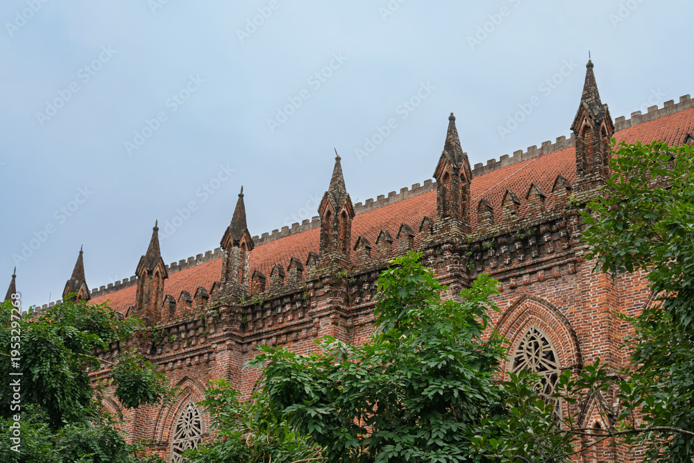Fototapeta premium Gothic brick facade of Chau Son Monastery with decorative arches and pointed spires partially covered by lush greenery under overcast sky.
