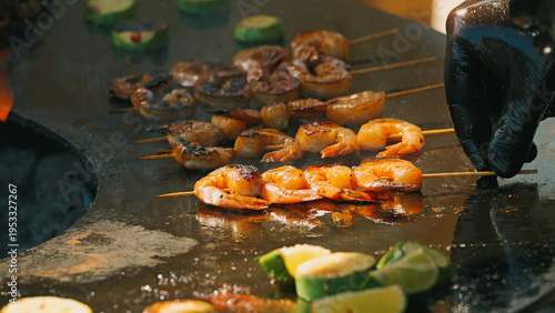 Close-up of a chef cooking seasoned shrimp skewers on a sizzling hot flat-top grill. Fresh lime and zucchini cook alongside. Appetizing street food concept at an outdoor market festival