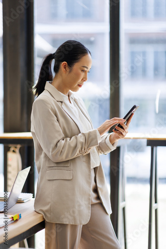 Confident Asian businesswoman using a smartphone while standing by a bright window in a modern office or cafe. Free Download Confident Asian businesswoman using a smartphone while standing by a bright window in a modern office or cafe. - GFXLoad