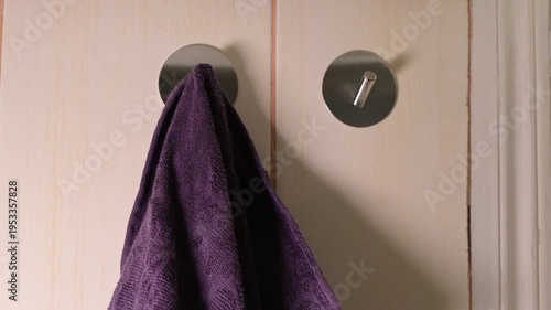 A man hangs two purple towels on metal hooks on the wall. Male hands alternately hang clean purple towels on round self-adhesive hooks fixed to light tiles in the bathroom