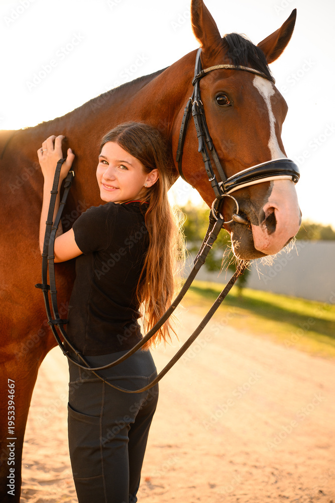 Obraz premium Smiling girl hugs horse while standing outside at sunset, looking happily at camera. Tender bond between rider and horse. Horse care concept.