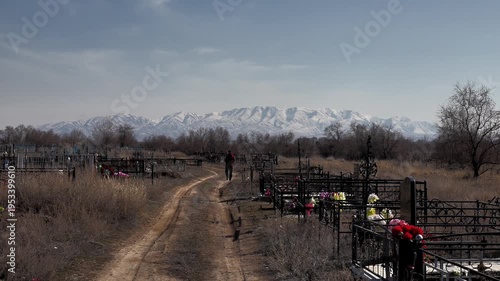 a man walks into the distance along a dirt road through an old rural cemetery against the background of graves and mountains in the snow in the background