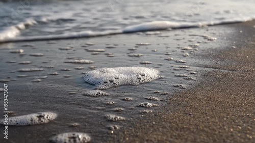 Ocean waves gently wash over sandy beach, creating foam and bubbles that shift and change as the tide recedes, capturing the serene beauty of coastal nature