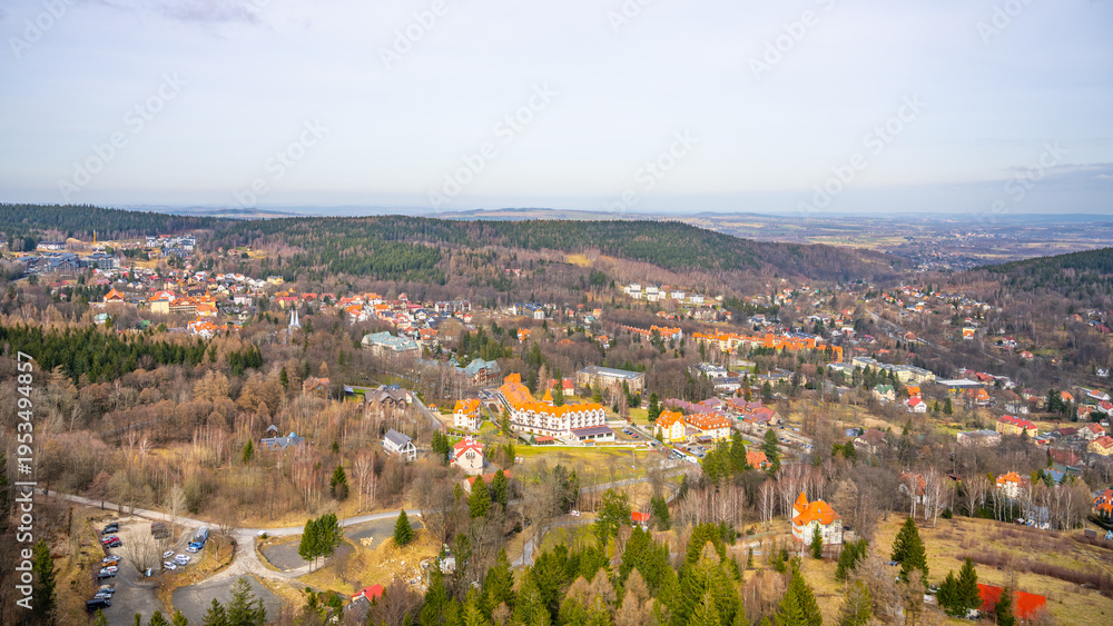 Fototapeta premium A wide view of a small town surrounded by trees and hills. Several buildings are visible, with roads and cars parked nearby. The scene shows a clear sky above.