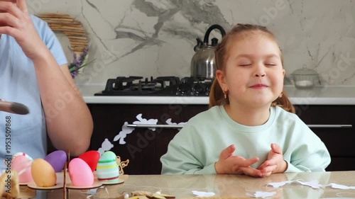 Mother and daughter are preparing for the Easter holiday together. A little girl is sitting at the kitchen table and waiting for a gift