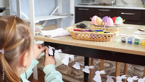 Back view of mother and her little cute daughter decorate the kitchen's table with a homemade paper garland. Festive preparations for Easter