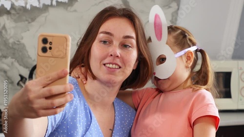 Portrait of happy mother and daughter in paper rabbit mask are preparing for Easter by taking selfies on smartphone together. Concept of Spring holiday on social media