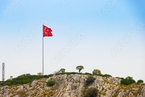 Obraz na płótnie Waving Turkish national flag on a high flagpole at the top of a steep rocky hill against a clear blue sky, panoramic view of a mountain summit for travel and national concepts