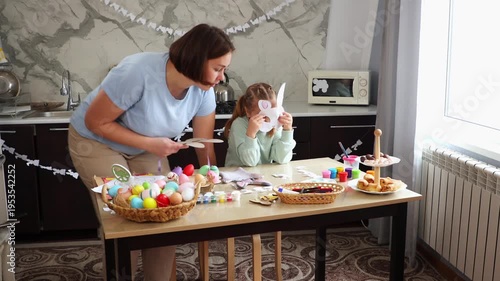 Wide shot of Caucasian mother and little daughter prepare for Easter by trying on a paper rabbit mask and playing together. Concept of Spring Festival
