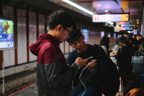 Two Young Asian Men Using Smartphone with Power Bank on Subway Platform