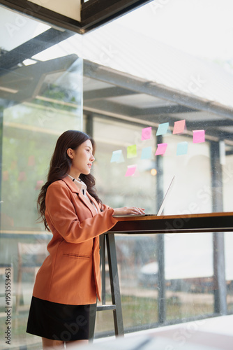 Young Asian businesswoman standing by a glass wall, typing on a laptop and collaborating on ideas with colorful sticky notes for business planning and strategy Free Download Young Asian businesswoman standing by a glass wall, typing on a laptop and collaborating on ideas with colorful sticky notes for business planning and strategy - GFXLoad