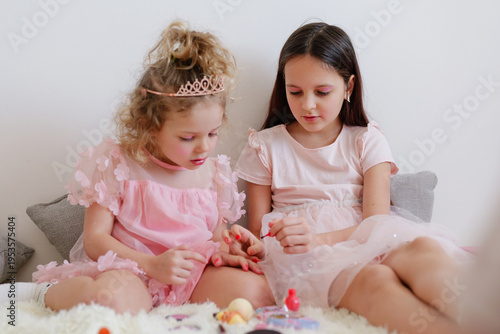 Two young girls sit on a soft surface at home applying nail polish to each other's nails. They appear focused and are enjoying their creative moment