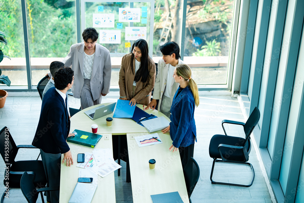 Fototapeta premium Business colleagues stand and discuss project details around meeting table in workspace while reviewing charts and documents. Concept of communication, teamwork and business planning.