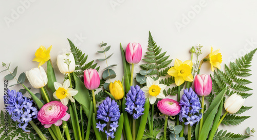 A beautiful and colorful border arrangement of various spring flowers including tulips, daffodils, hyacinths, and ranunculus with green foliage on a light background.