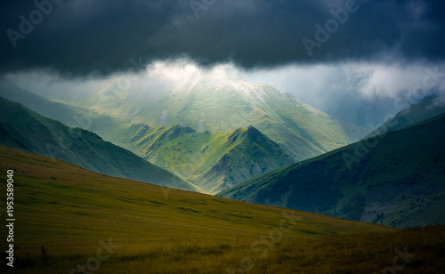 Dramatic mountain landscape with sunlit green peaks beneath dark storm clouds. Dramatic natural scenery with layered ridges, moody weather, and striking contrast in a remote highland setting