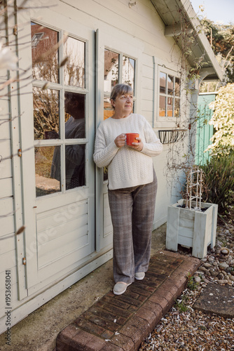 Mature woman standing by house holding mug in garden sunlight
