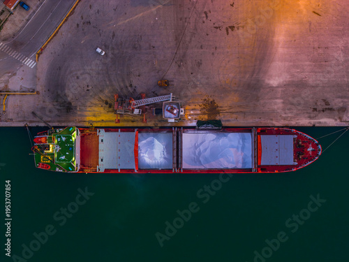 Aerial view of a cargo ship docked at the port, its red hull contrasting with the deep green water and the pale dockside, Vasto, Abruzzo, Italy.