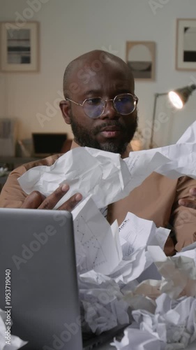 Vertical shot of stressed African American man covering face with hand while surrounded by messy crumpled documents on office desk