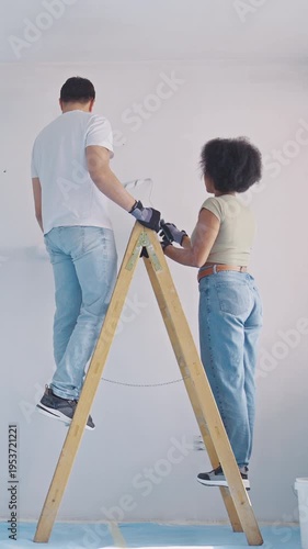 A young couple collaborates on a home renovation project. The hispanic man paints a wall while the african american woman steadies the ladder.