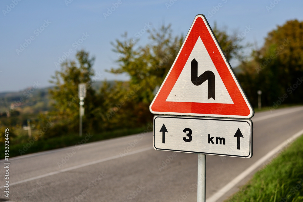 Obraz premium Close-up of a triangular road warning sign indicating winding curves ahead for the next 3 kilometers, set along a rural asphalt road in the Province of Siena, Italy.