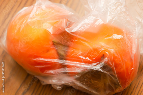 Ripe orange persimmons are inside a transparent plastic bag on a wooden table surface. The image illustrates a common household situation of storing products or buying fruit, highlighting the texture 