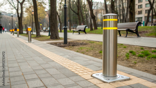 Stainless steel security bollard with a yellow reflective stripe on a modern city sidewalk near a park, urban safety infrastructure. Street protection, city planning.