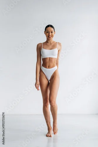 Obraz Slim female model wearing white bikini poses confidently in a bright studio with a clean, minimalistic background emphasizing her physique and fashion style