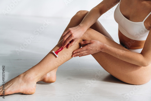 Female body in white underwear using a razor to shave her leg while seated on a wooden floor in a bright indoor setting