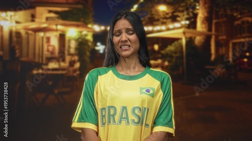 Young brunette woman in yellow brazil jersey grimacing, face contorted under cafe lights on street; unease.