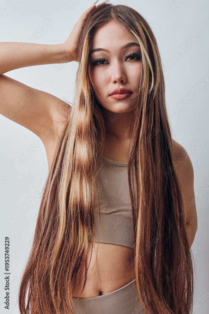 Obraz premium Young woman with long straight hair wearing beige crop top posing in studio. Beauty portrait with natural makeup, confident expression and soft lighting on white background.