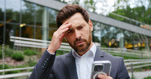 Quadro su tela Close-up of a concerned bearded businessman in a business suit, holding a smartphone and reacting emotionally to an urgent message outdoors in a city park