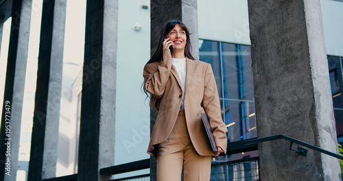Obraz na plátne Confident businesswoman in a formal suit holding a laptop and talking on the phone while walking outdoors