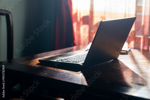 A laptop and spiral notebook on a wooden table with soft morning sunlight. Concept of remote work, home office, and professional productivity.

