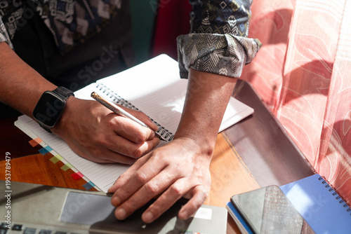 High angle shot of a person wearing batik shirt writing notes in a spiral notebook next to a laptop and smartphone on a wooden desk with sunlight.

