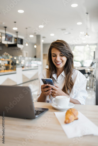 A happy young woman checks her smartphone while sitting at a table with a laptop, coffee, and croissant in a bright modern cafe.