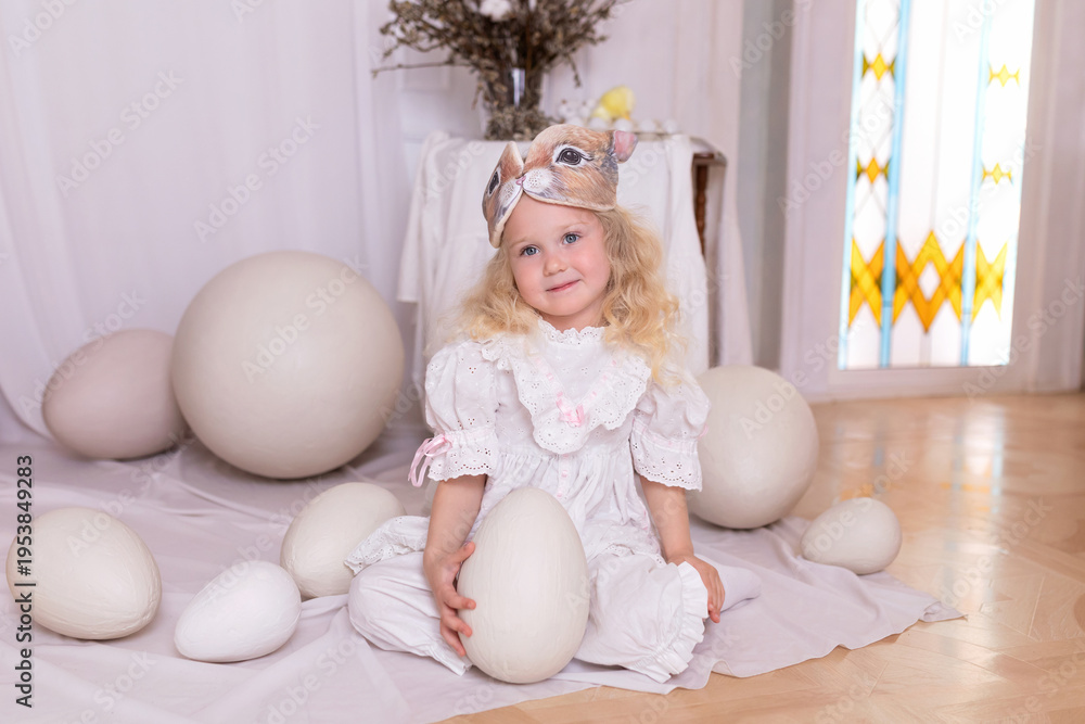 Fototapeta premium A little blonde girl in a white dress and a rabbit ears mask on the head is smiling sit with with large Easter eggs. Happy child girl with bunny ears holding an easter egg in her hands. Vintage style