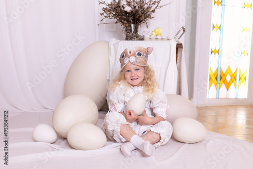 Wallpaper Mural A little blonde girl in a white dress and a rabbit ears mask on the head is smiling sit with with large Easter eggs. Happy child girl with bunny ears holding an easter egg in her hands. Vintage style Torontodigital.ca