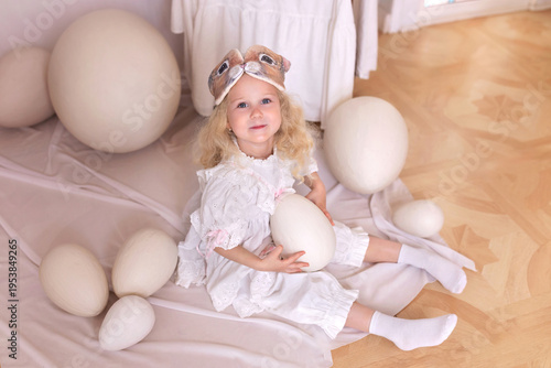Wallpaper Mural A little blonde girl in a white dress and a rabbit ears mask on the head is smiling sit with with large Easter eggs. Happy child girl with bunny ears holding an easter egg in her hands. Vintage style Torontodigital.ca