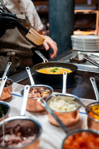 A chef sautes an omelet in a black skillet, adds herbs, and plates by copper pans with onions, peppers, tomatoes, and mushrooms in a warm, upscale Zermatt hotel kitchen.