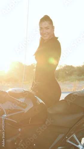Lovely cheerful 25-aged trendy woman with high ponytail dresses in black wear standing near her sporty black motorcycle on car wash and looking at camera with sincerely smile,close up