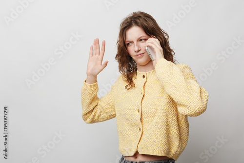 Young woman in yellow sweater talking on phone and waving hand. Casual communication concept with friendly gesture on light background. Modern lifestyle and social interaction.