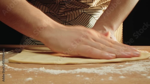 Hands of caucasian man pressing dough into round on floured table. Fast rhythmic motion with rustic apron and warm kitchen light. Shaping thin puff pastry for pizza baking in oven by artisan
