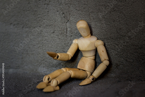 A wooden mannequin sits against a gray background, leaning against a wall. Its pose suggests weariness or thoughtfulness, creating an atmosphere of loneliness and sadness.
