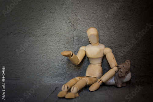 A wooden mannequin sits against a gray wall, its hand outstretched as if begging. A small dog figurine crouched beside it—a faithful companion in solitude.