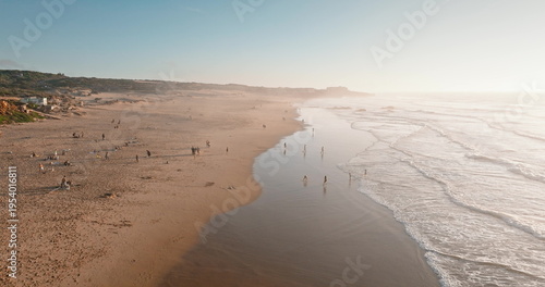 Portugal, Lisbon: Praia do Guincho beach coastline at sunset, people relaxing on sand, walking along the water edge with gentle ocean waves rolling under clear sky. Aerial drone flight