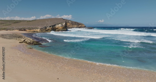 Penisok beach in Lombok, Indonesia, featuring a rugged coastline with cliffs, golden sand, and turquoise ocean water crashing with white waves under a clear blue sky. Travel vacation background