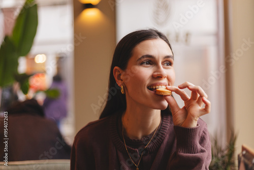 Cuadro en lienzo Happy and cheerful young woman eating a small cookie or sweet snack while sittin