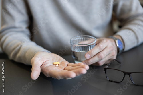 Foto Senior man holding daily pills in his palm with a glass of water nearby, taking
